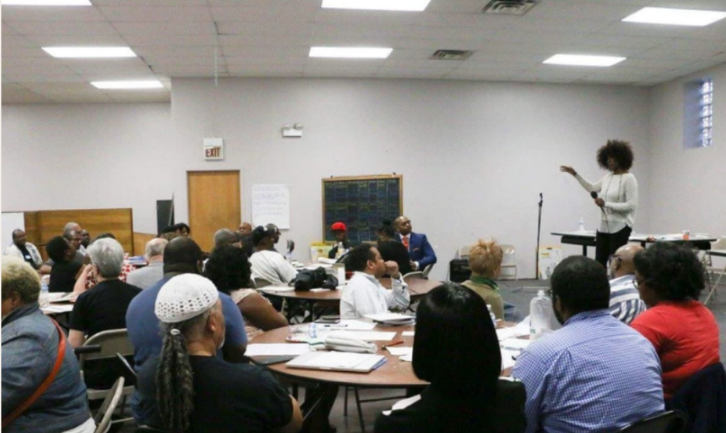Residents seated around tables during a community meeting, with a speaker at the front.