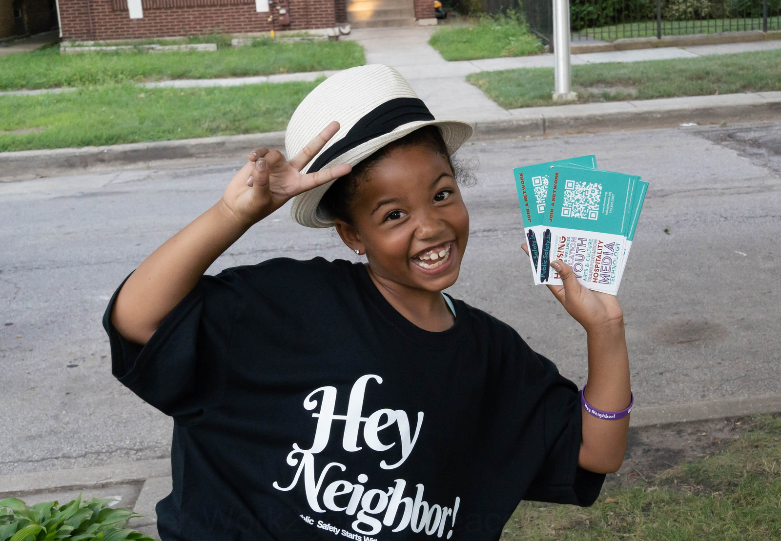 A smiling young participant in a white hat and Hey Neighbor shirt holding neighborhood information